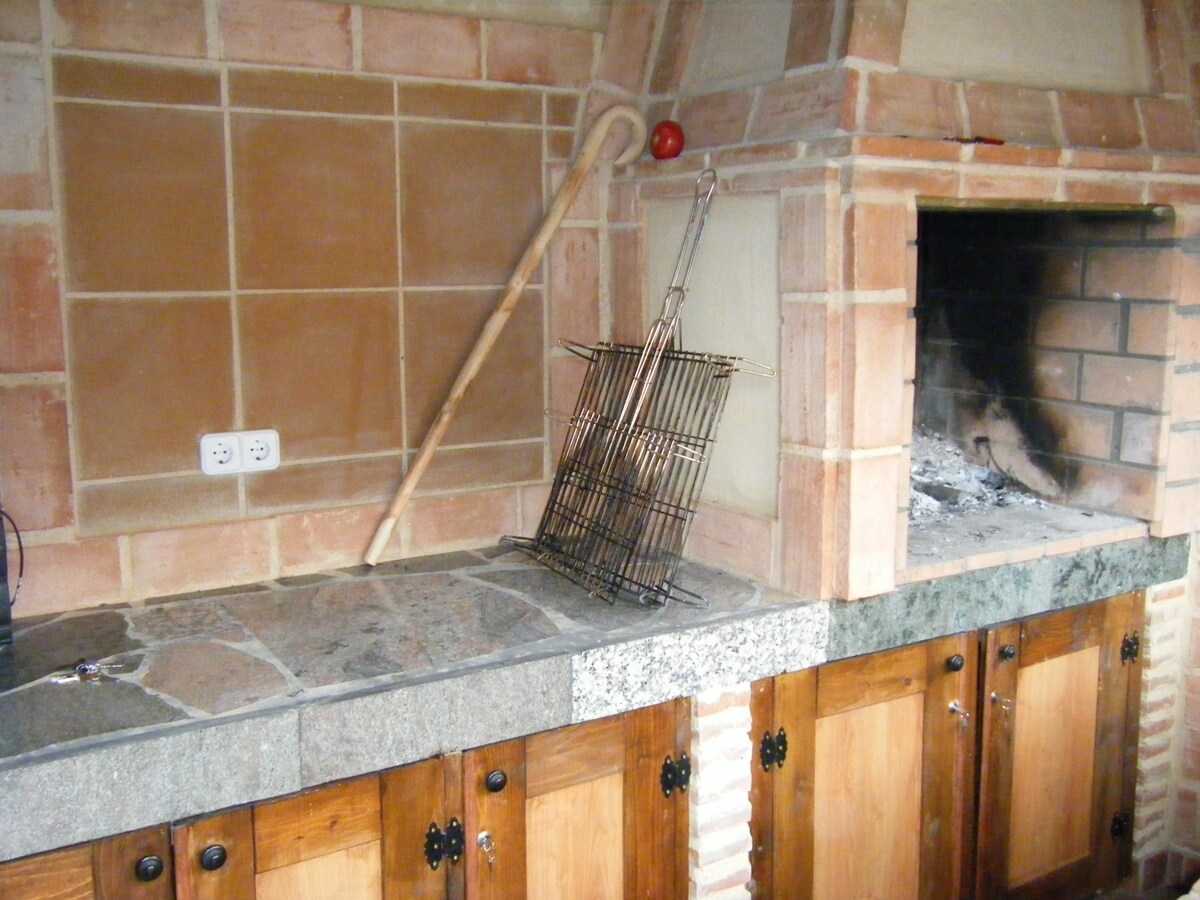 An outdoor barbecue area is presented, featuring a stone countertop with a metal grill and a wooden handle resting against the wall. The grill is positioned next to a brick fireplace with remnants of ashes. Below, wooden cabinets provide additional storage space.