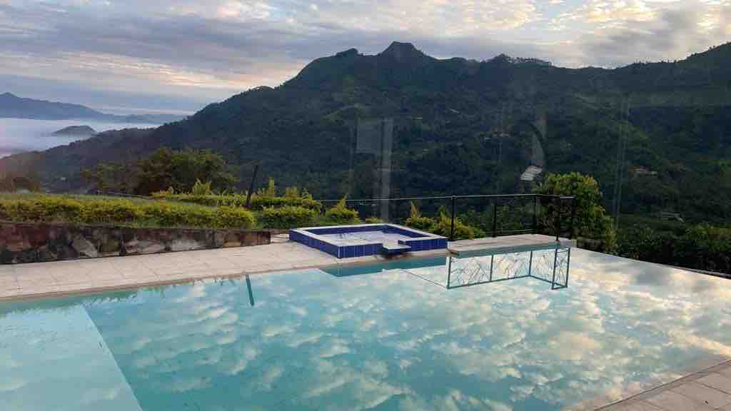 A serene outdoor area features a clear pool reflecting the sky, surrounded by green hills. A spa located near the pool adds an additional relaxation option. The landscape is characterized by lush vegetation and distant mountains under a cloudy sky.
