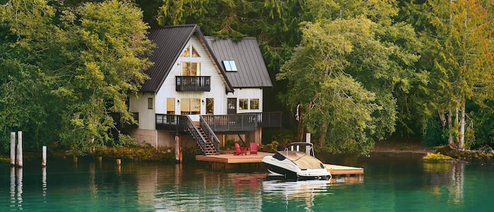 Une photo montre un bateau, couvert et amarré devant une maison de 2 étages située au bord d'un lac.