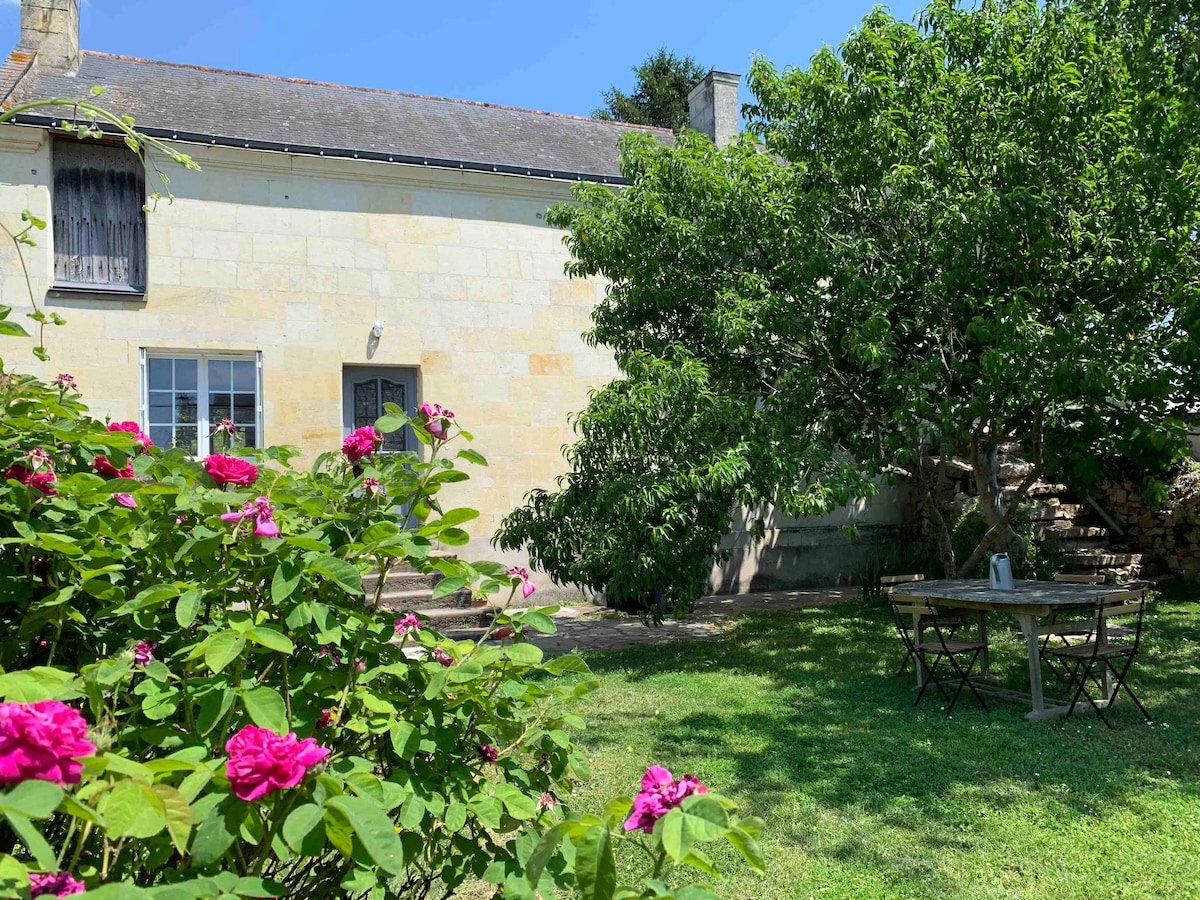 The exterior of a light-colored house is surrounded by green grass and vibrant flowering bushes. A wooden table with two chairs is positioned under a leafy tree, providing a shaded area for relaxation. The house features a small set of stairs leading up to the entrance.
