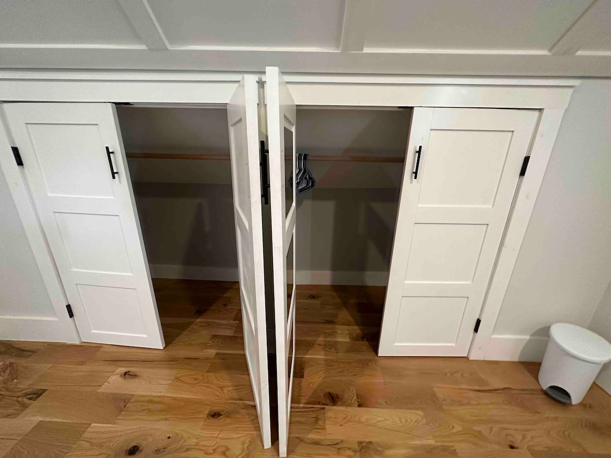A set of white pocket doors stands open, revealing a spacious closet with light wooden flooring. Hangers are neatly arranged on the upper rail, while a small wastebasket is positioned beside the closet. The clean lines of the doors complement the overall design.