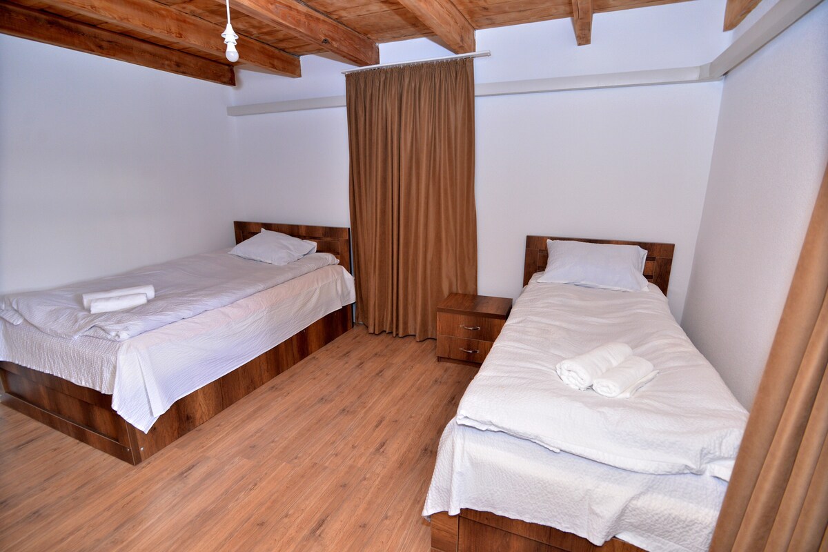 A simple bedroom is seen, featuring two wooden beds with light bedding. A nightstand is located beside one bed, and a curtain separates the space from another area. Natural light enters from an adjacent window, illuminating the wooden floor.