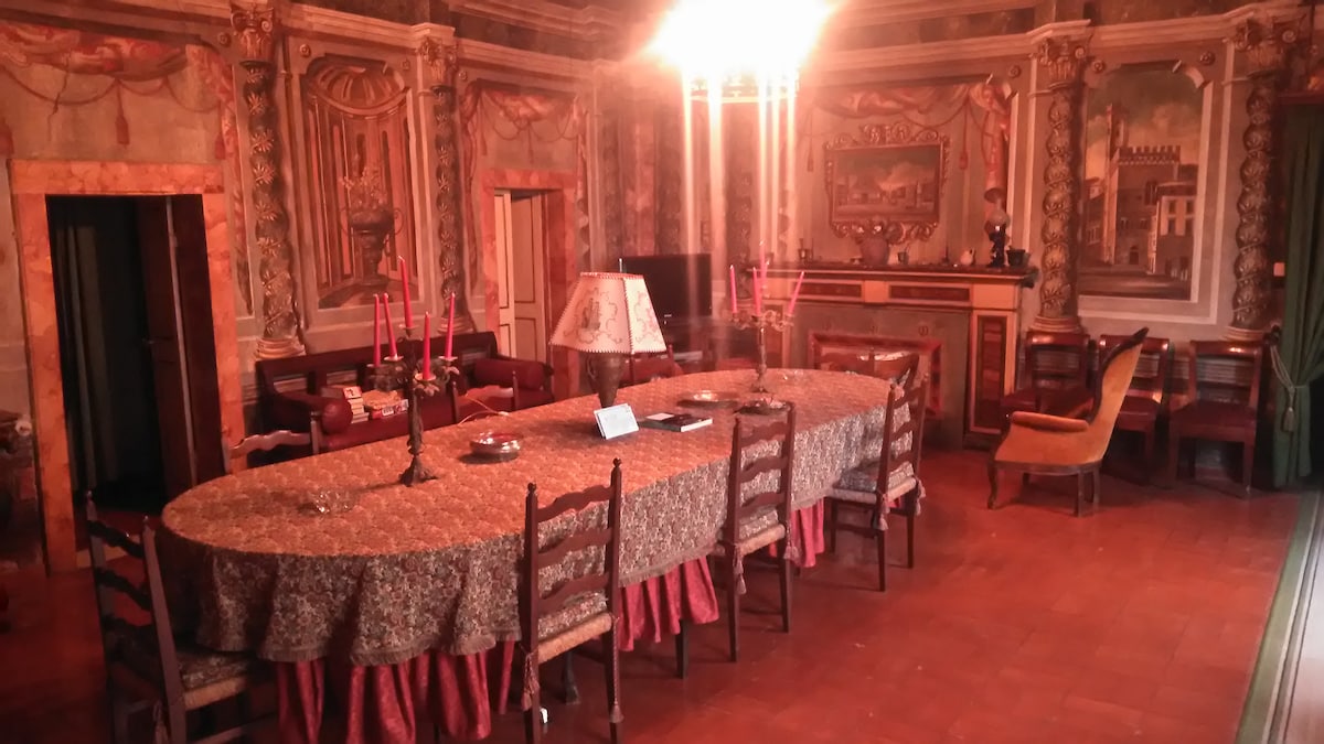 An elegantly furnished dining room is featured, highlighting a long, oval table surrounded by wooden chairs. Intricate wall murals and decorative lighting create a refined ambiance. A vintage lamp sits on the table, complemented by candles and decorative items along the table's center.