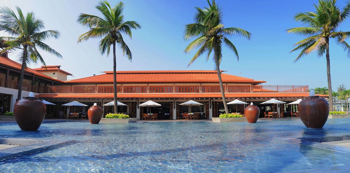 The expansive pool area features a stunning outdoor dining space, surrounded by palm trees. Umbrellas provide shade for the tables, while large clay pots add a touch of elegance. The villa's architecture harmonizes with the tropical landscape, reflecting a serene environment.
