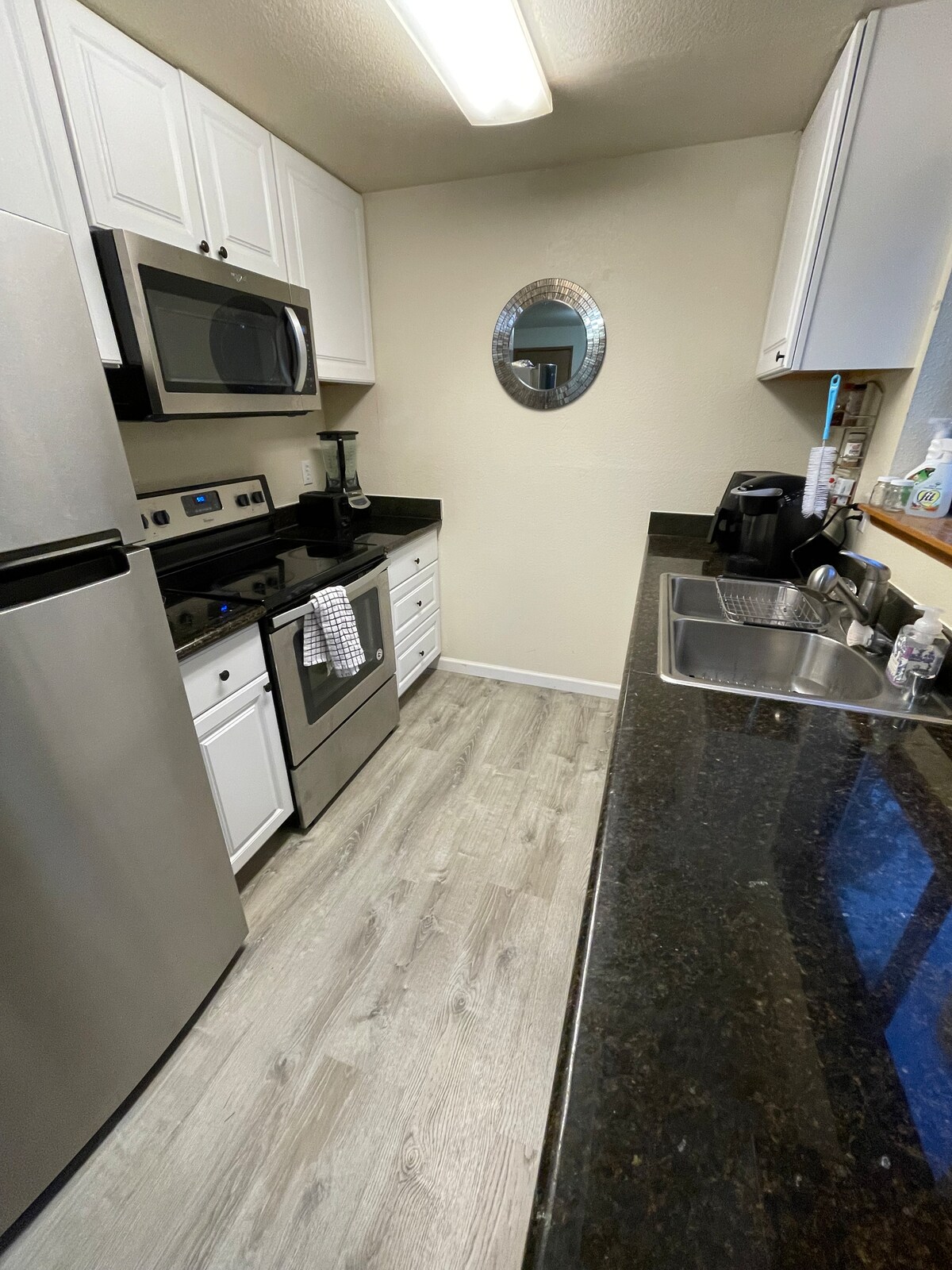 A modern kitchen is equipped with stainless steel appliances, including a refrigerator, microwave, and oven. White cabinets and dark countertops create a clean contrast. A round mirror adds a touch of design, while ample counter space is available for meal preparation.