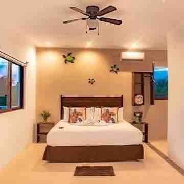 A well-lit bedroom features a spacious bed adorned with decorative pillows. Light-colored walls complement dark wooden furnishings, including bedside tables. A ceiling fan contributes to the airy atmosphere, while windows allow natural light to brighten the room.