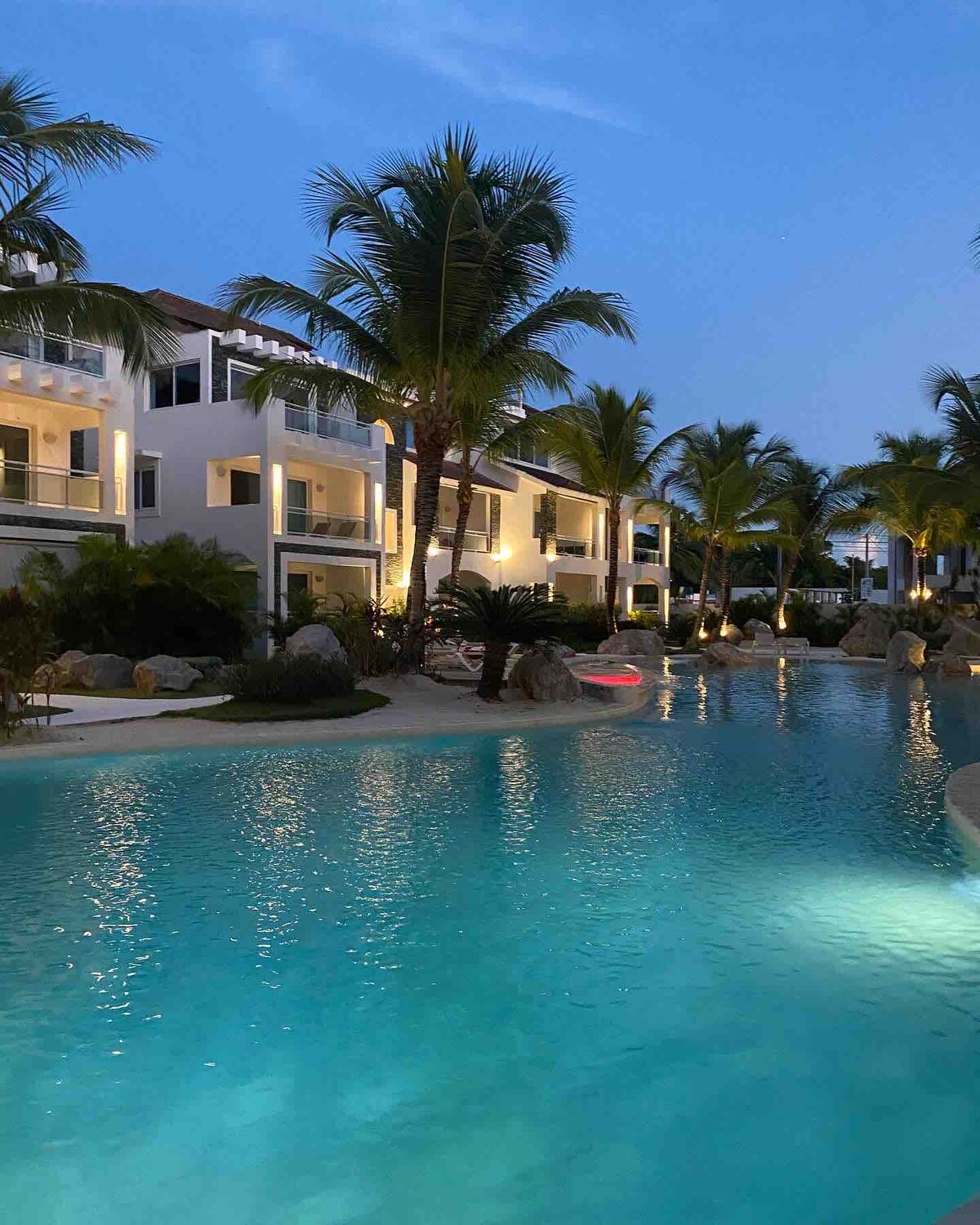 The image captures a serene evening view of a pool area surrounded by lush palm trees. Soft lighting accentuates the curves of the water, while modern buildings peek through the greenery, creating an inviting atmosphere for relaxation.