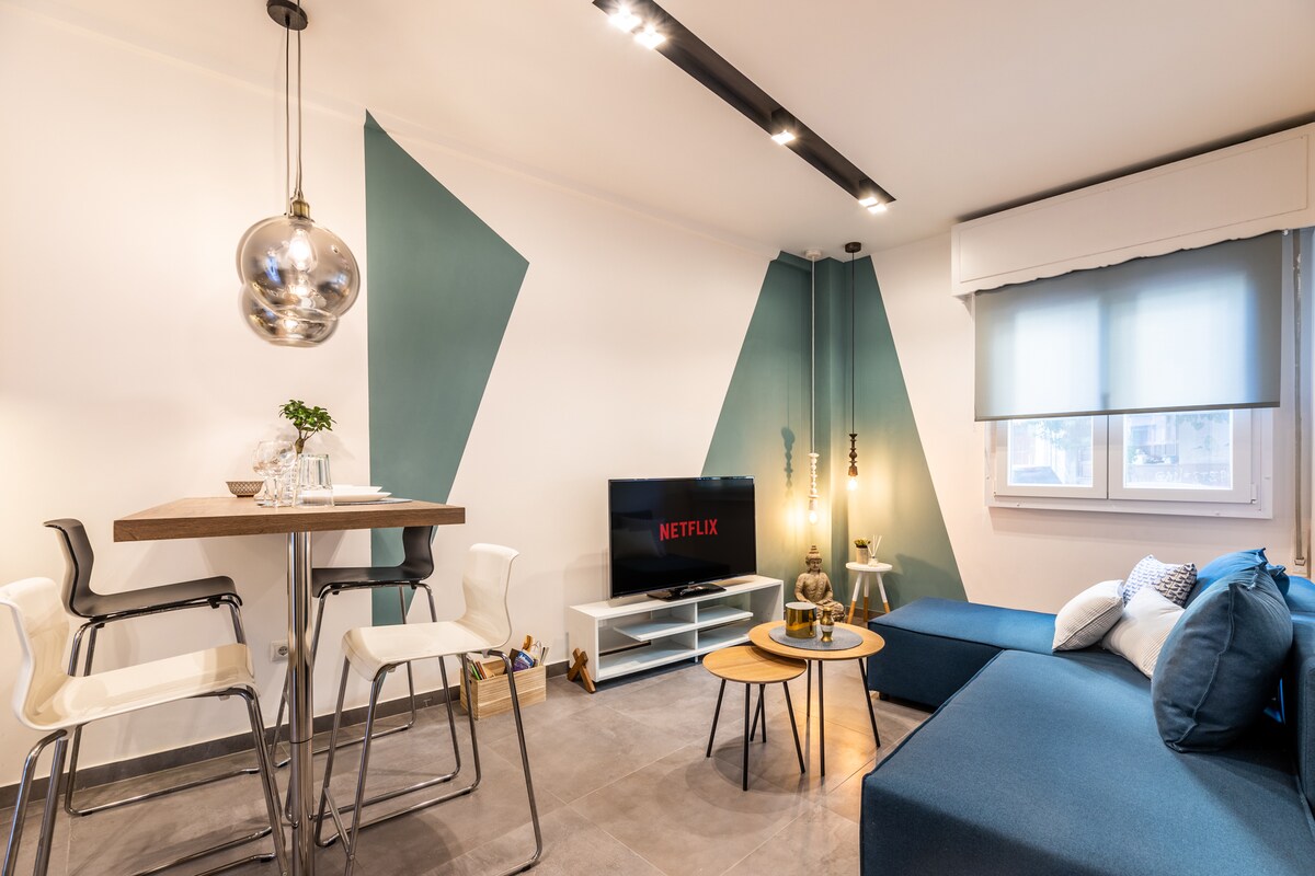 A stylish living area features a comfortable blue sofa and a pair of minimalist wooden tables. A television mounted on the wall displays the Netflix logo. A modern lamp and decorative items add a touch of personality. Natural light filters through the window, enhanced by light-colored shades.