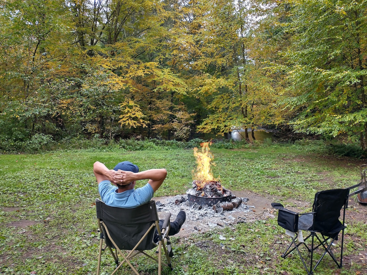 A seating area features two camping chairs arranged around a fire pit, which displays a vibrant flame. The surrounding landscape showcases lush green grass and trees displaying autumn foliage, with a stream visible in the background.