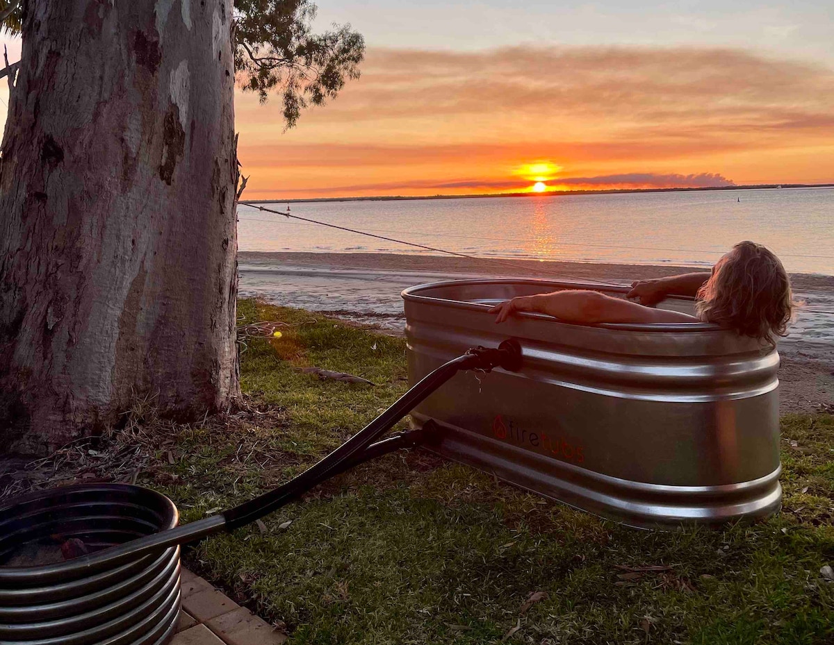 A hot tub is positioned under a tree, with a person relaxing inside. The vibrant sunset casts warm hues over the tranquil waters, creating a serene atmosphere. The shoreline and distant horizon are visible in the background, enhancing the picturesque coastal setting.