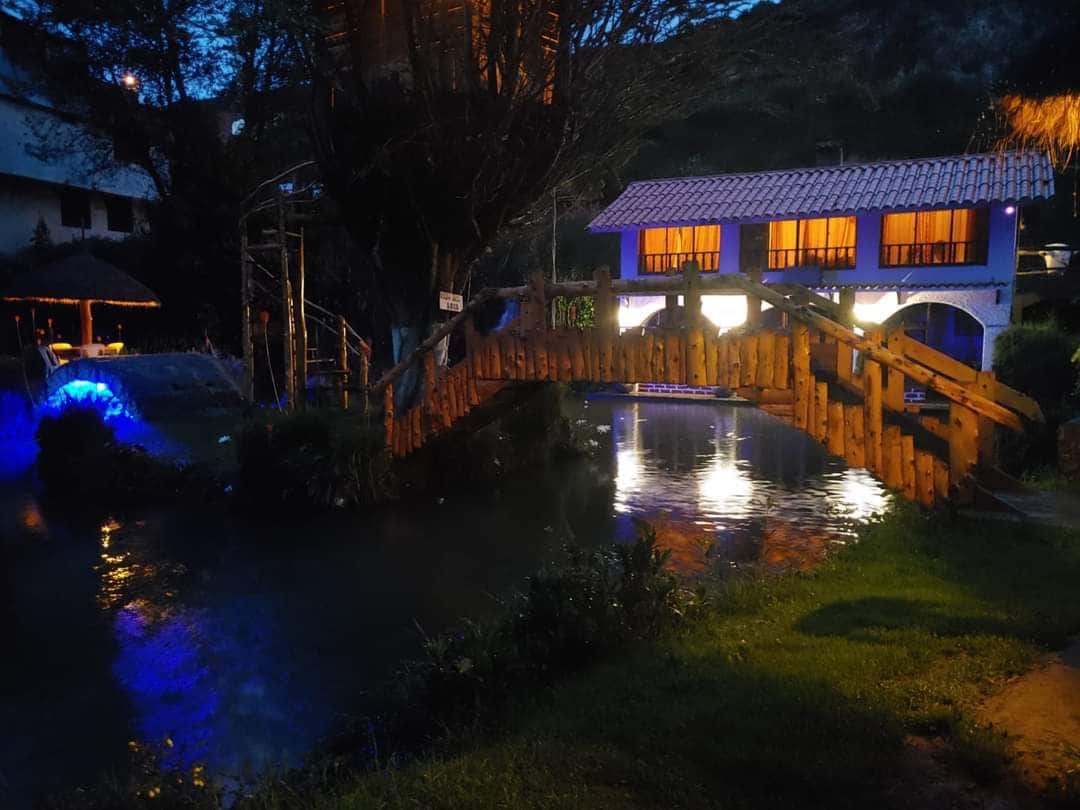 A charming wooden bridge extends over a serene body of water, reflecting soft blue and warm yellow lights from nearby buildings. The surrounding landscape features lush greenery, and the night sky adds a peaceful ambience to the scene.