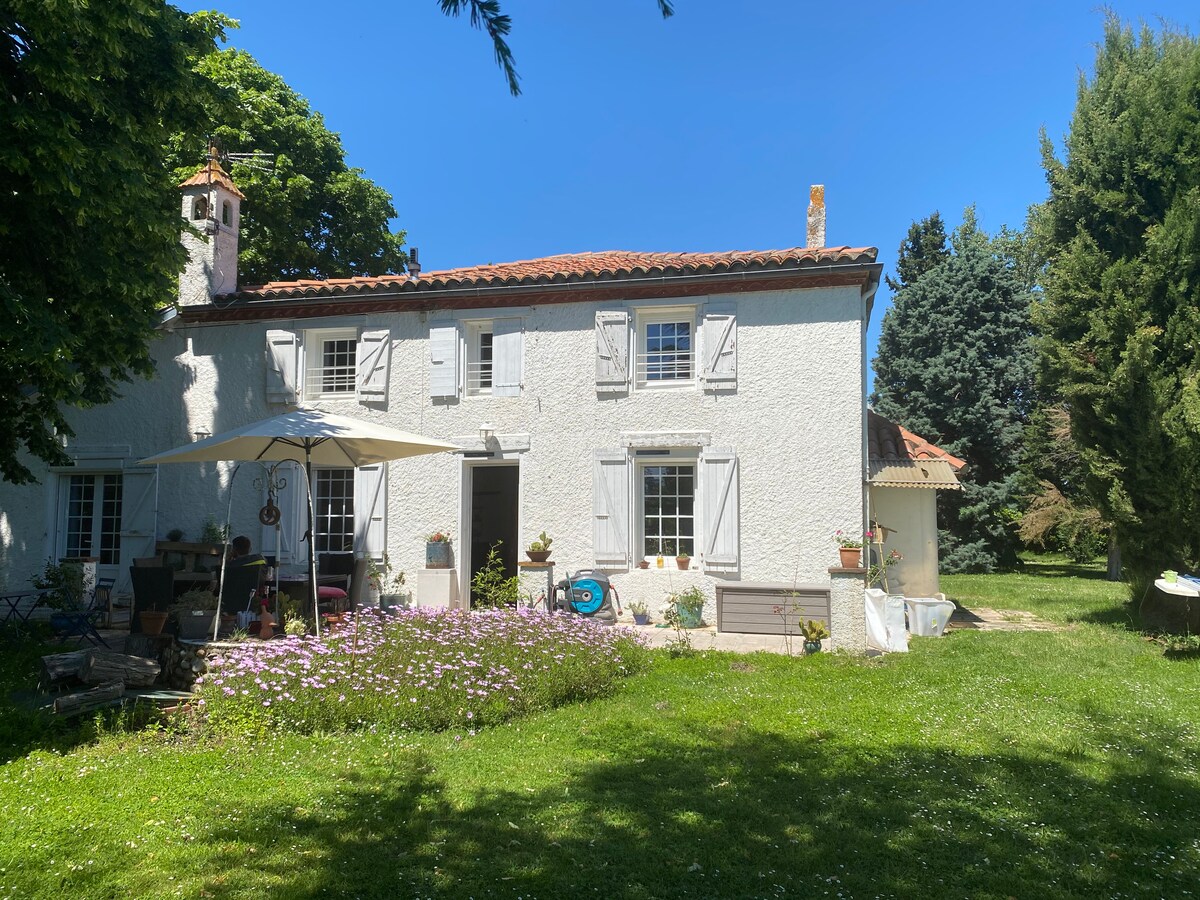 A traditional house is presented in a serene setting, featuring white exterior walls and grey shutters. A patio area offers an umbrella over outdoor seating among flowering plants, leading to a spacious green lawn bordered by tall trees.