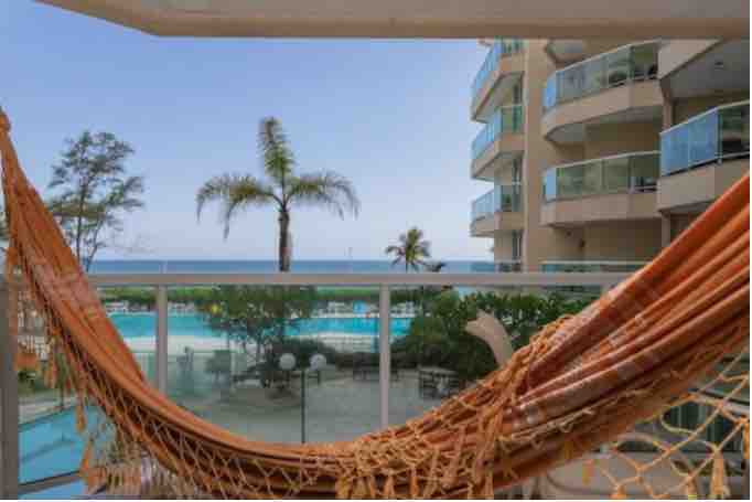 A balcony view features a relaxed hammock, framed by a tranquil setting that includes a pool and ocean. Palm trees line the stunning landscape, enhancing the serene atmosphere. The sunlight bathes the area, inviting peaceful moments in a picturesque environment.
