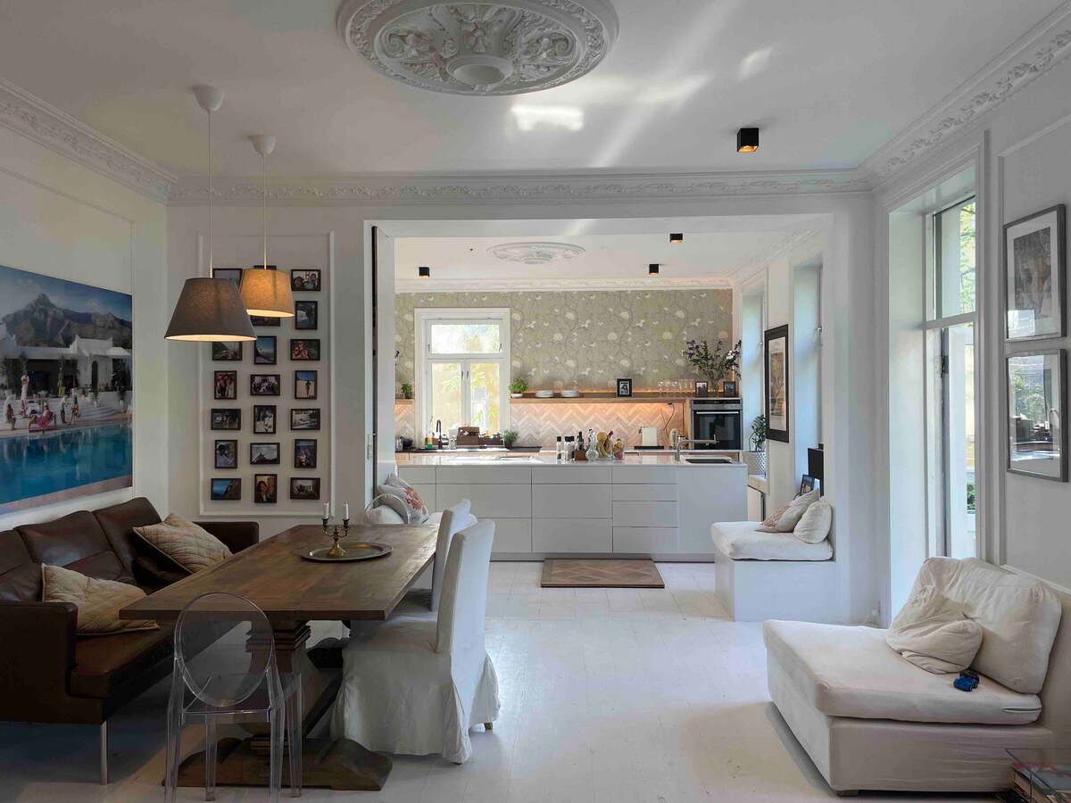 A spacious living area features a large dining table surrounded by chairs and a comfortable sofa. Natural light enters through multiple windows, illuminating the classic ceiling details and contemporary décor. In the background, an open kitchen is visible, showcasing modern appliances and stylish finishes.
