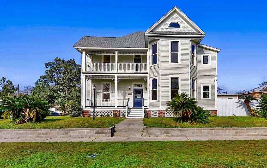 A charming two-story house features a light-colored exterior and a front porch with white railings. Lush palm trees accent the well-maintained yard, and large windows allow for ample natural light. The entrance is framed by decorative architectural details, enhancing the inviting appearance.