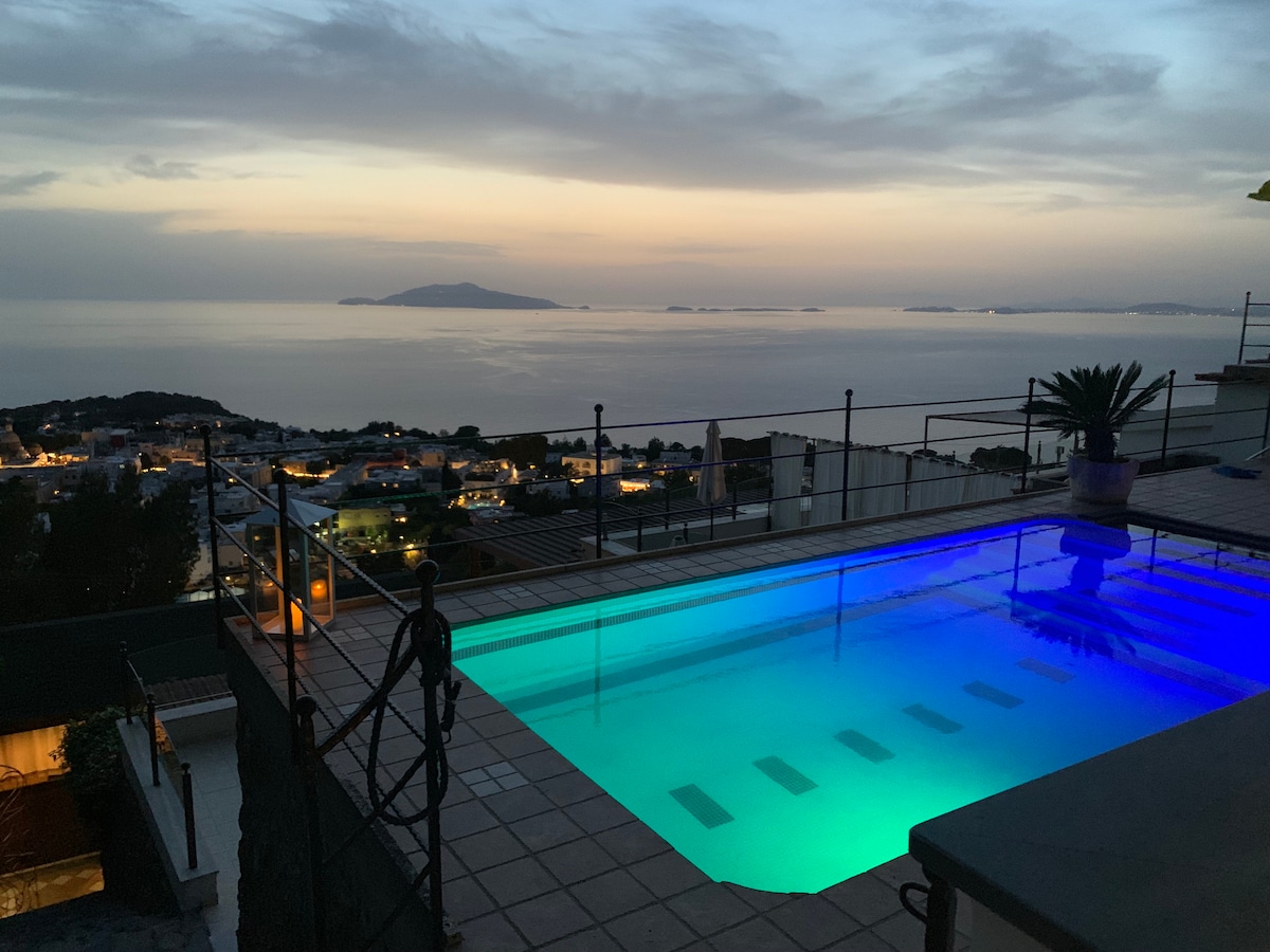 An outdoor pool is illuminated with a calming blue light, situated against a backdrop of a sunset over the Mediterranean Sea. The surrounding area features tiled surfaces and a railing, with distant lights from the nearby village creating a serene atmosphere.