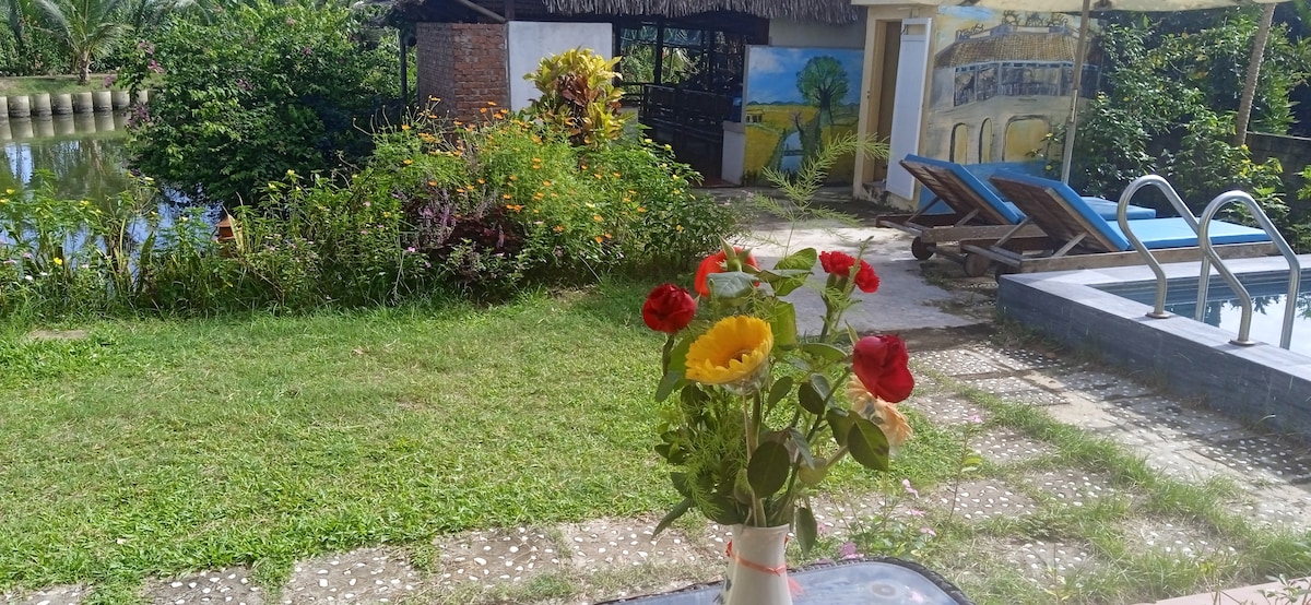A vase containing bright flowers is placed on a table, with a scenic view of a grassy area leading to a swimming pool. Lush greenery surrounds the space, while a charming pavilion can be seen in the background, complementing the tranquil environment.