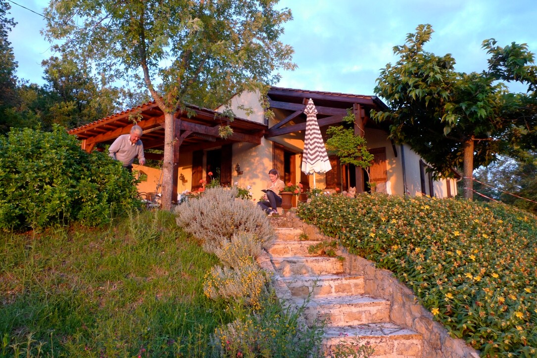 The house is set on a sloping ground, surrounded by lush greenery and blooming flowers. Stone steps lead to a welcoming entrance, with an umbrella-covered table and outdoor seating creating an inviting space for relaxation.