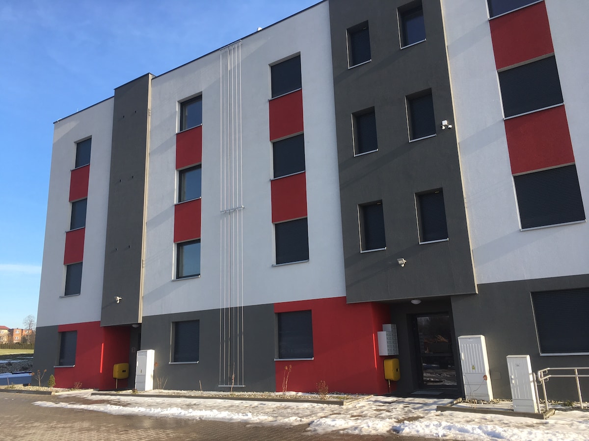 The exterior of the modern building features a combination of grey, white, and red panels. The façade includes multiple windows with dark coverings and an entrance marked by a well-defined doorway. Snow is visible on the ground, indicating the season.