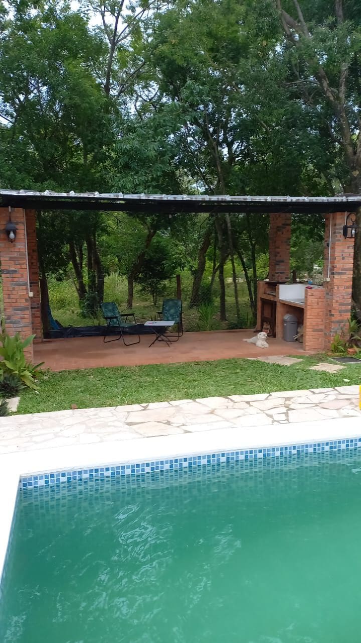 A shaded outdoor seating area is visible, featuring two green chairs and a small table under a covered structure. Beyond, lush greenery surrounds the space, and a sparkling pool is in the foreground, framed by a stone border.