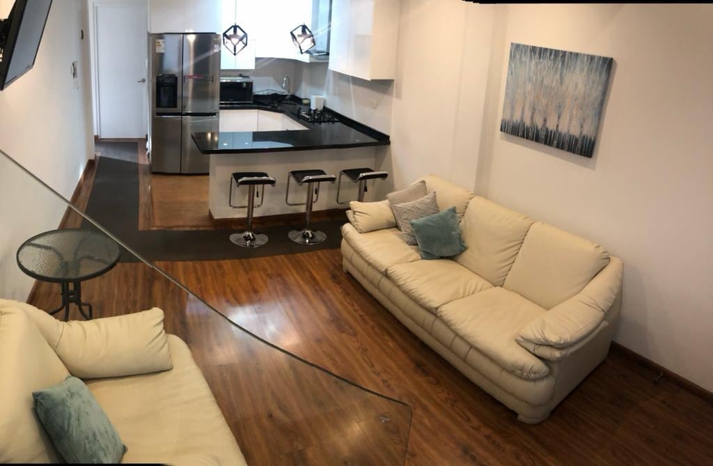 A modern living area features two light-colored sofas and a glass coffee table. The kitchenette is visible in the background, equipped with stainless steel appliances. Bar stools are arranged at the counter, and a piece of abstract art adorns the wall.