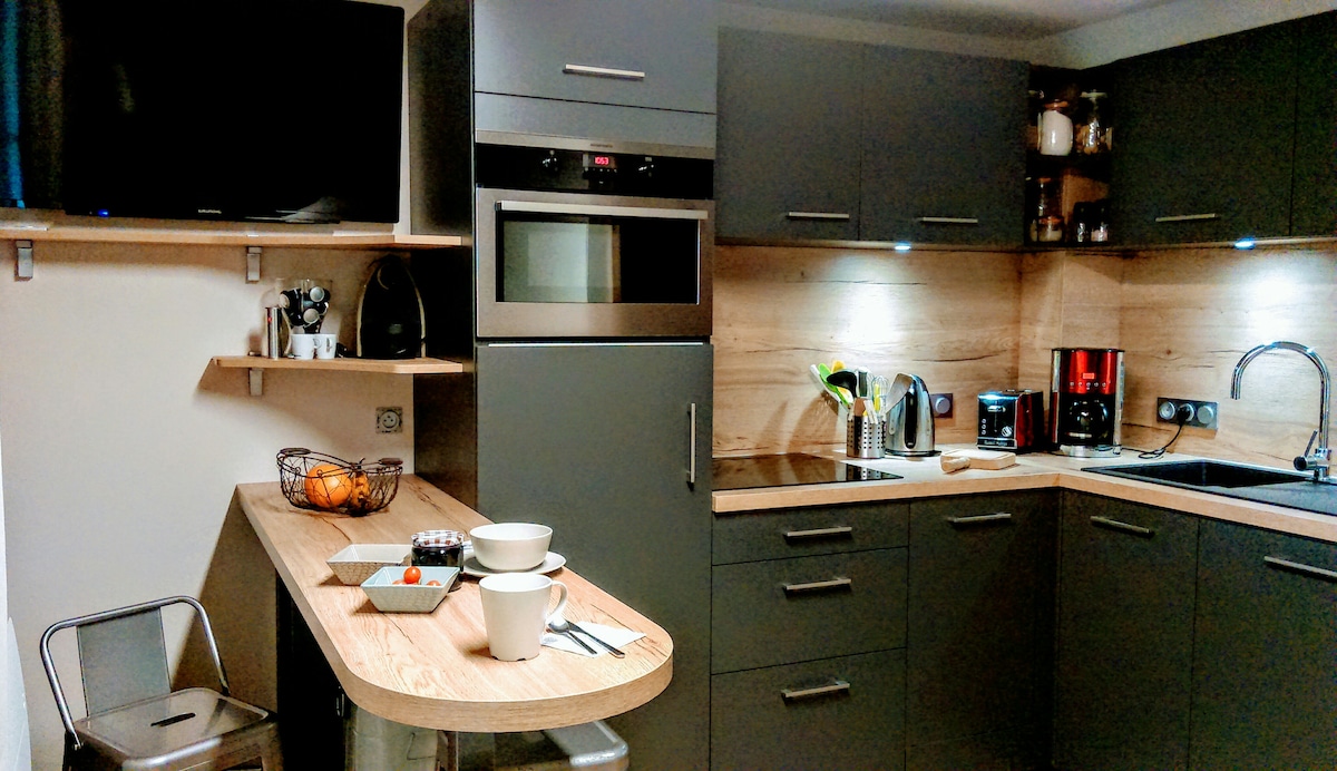 A modern kitchen is presented, featuring sleek grey cabinetry and wooden accents. Equipped with built-in appliances, it includes a microwave, oven, and coffee maker. A small table with bowls and utensils is situated nearby, with ample counter space for meal preparation.
