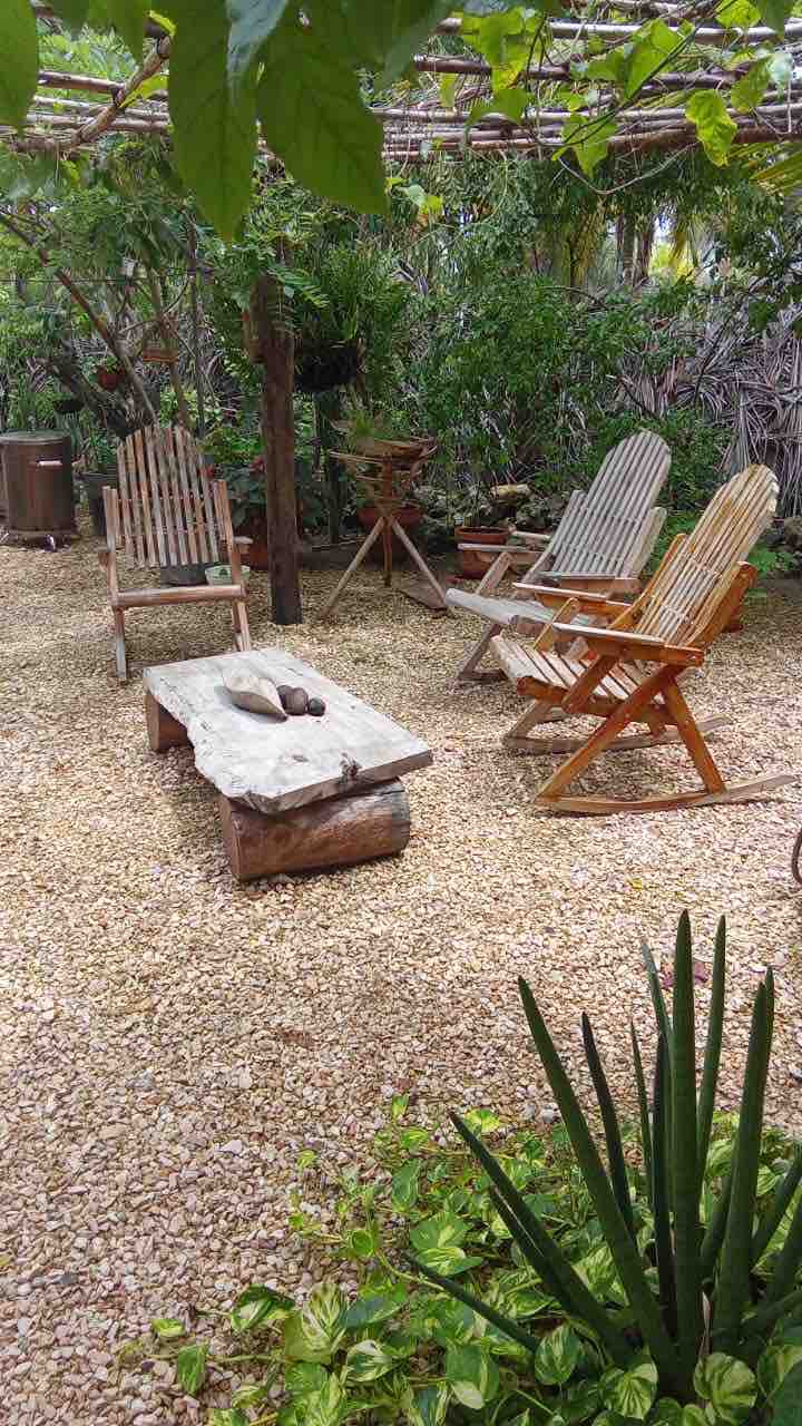 A tranquil outdoor space is presented, featuring wooden adirondack chairs arranged around a natural stone coffee table. Lush greenery surrounds the area, with various plants and trees providing a serene backdrop. The ground is covered with small gravel stones, enhancing the peaceful ambiance.