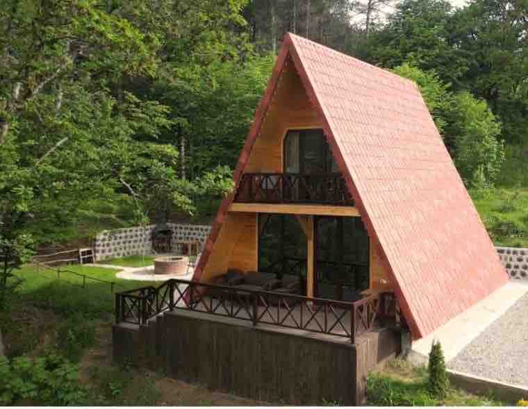 A distinctive A-frame cabin is surrounded by lush greenery. Large glass windows allow natural light to fill the interior. Two balconies provide outdoor seating areas, and a stone fire pit is visible in the grassy yard, inviting relaxation in a serene setting.