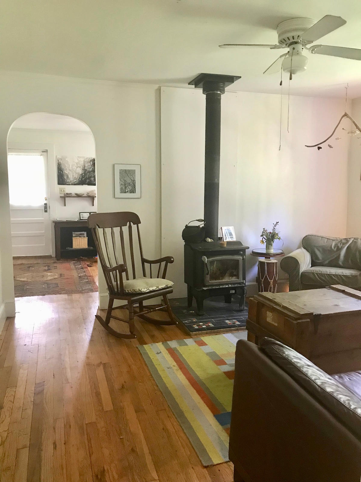 A light-filled living area showcases a wooden rocking chair beside a wood-burning stove, enhancing the room's rustic charm. A green sofa and a wooden trunk add comfort. An area rug with colorful stripes defines the space, while doorways lead to other parts of the home.
