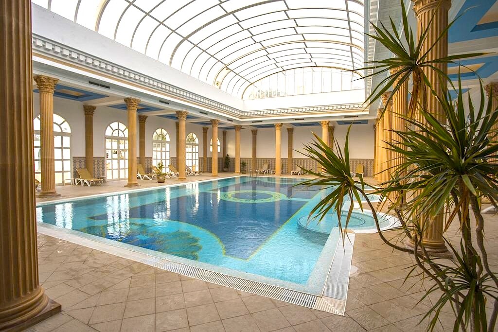 An indoor swimming pool is surrounded by elegant columns and features a modern glass ceiling. Lounge chairs are positioned along the pool's edge, alongside decorative tiles in varying shades of blue. Natural light illuminates the space, creating a serene swimming environment.