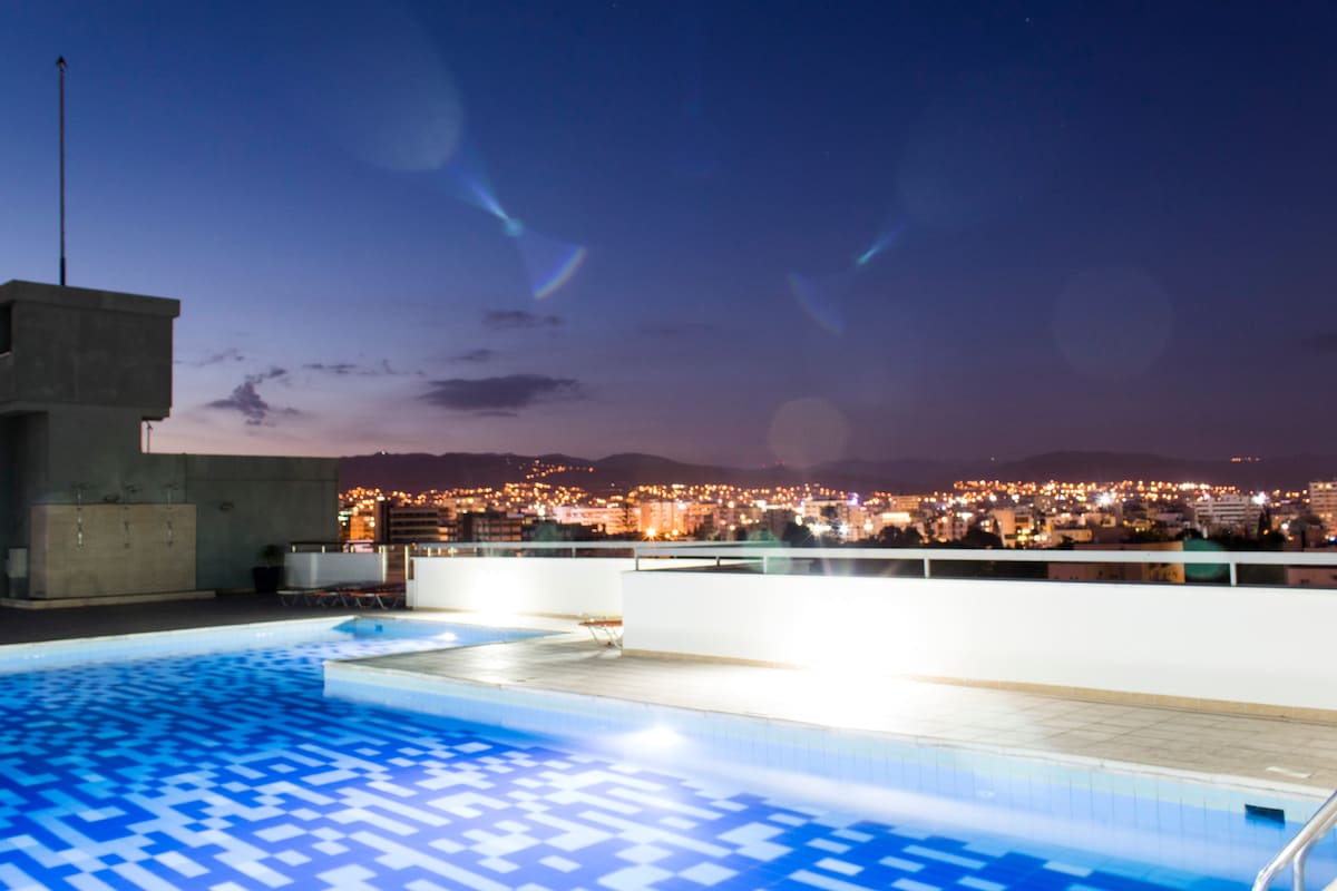 The rooftop area features a swimming pool with a geometric blue tile pattern. Nighttime views of the city are illuminated by distant lights. A clear sky is visible above, while the pool's edge reflects the surrounding ambiance.