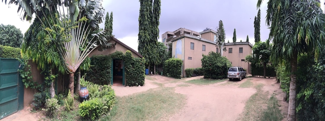 The exterior of the property features a driveway leading to a landscaped area surrounded by tall trees and lush greenery. Buildings with neutral-colored walls are visible, along with a parked vehicle. The scene is set under a partly cloudy sky.