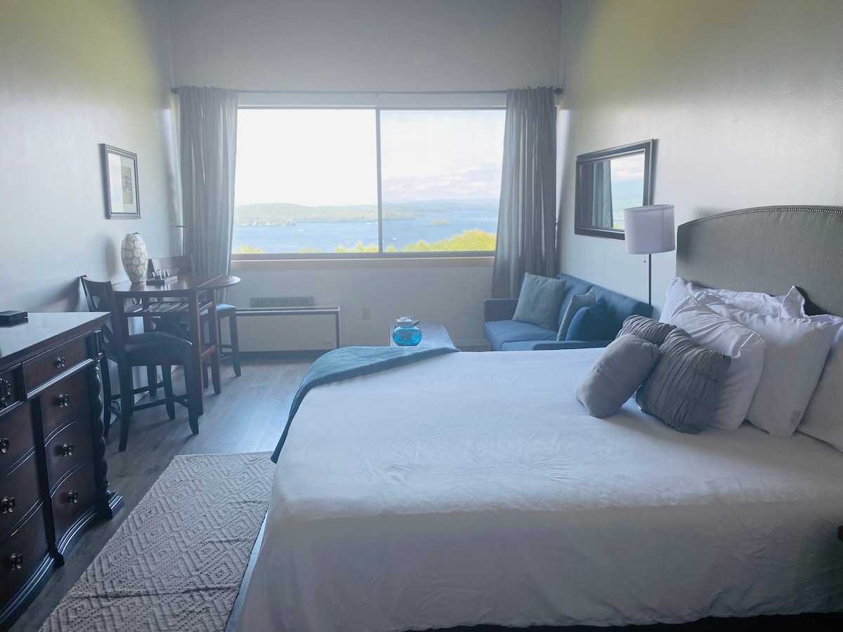 A spacious studio layout features a large king-size bed adorned with decorative pillows. A seating area with a couch is present, and a dining nook is positioned by the window, allowing for lake views. Light curtains frame the expansive window showcasing the scenery.