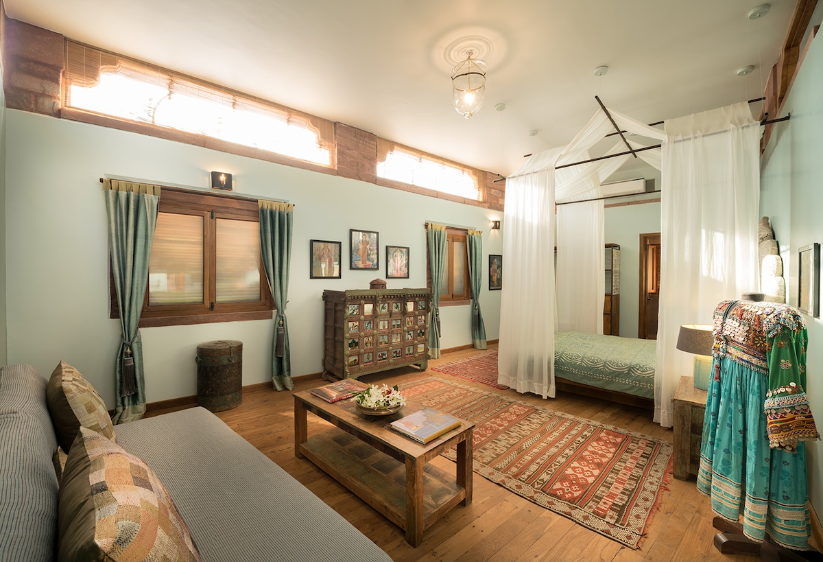 A spacious room features a double bed draped with a light canopy, complemented by a comfortable sitting area with a gray sofa and a wooden coffee table. Vintage textiles and artwork adorn the walls, while large windows allow natural light to fill the room.
