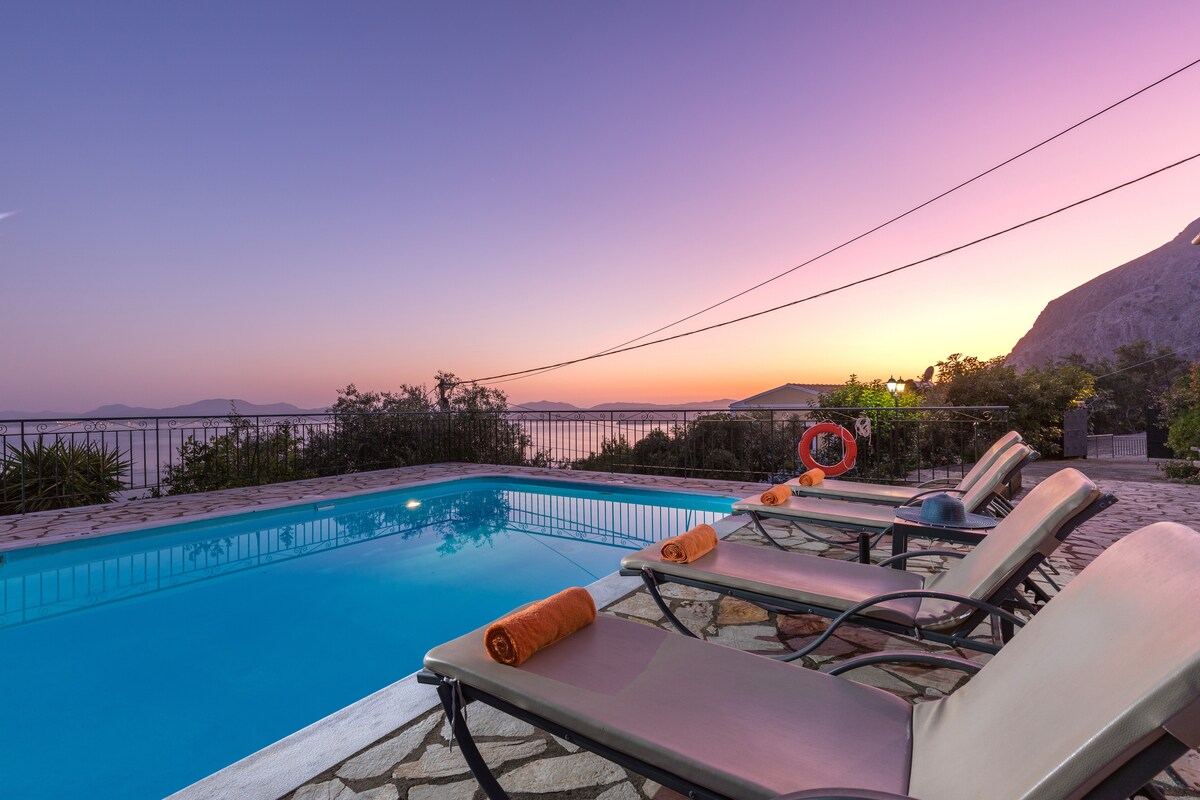 Loungers with orange towels are positioned alongside a private swimming pool, reflecting the serene hues of a sunset sky. The surrounding area showcases natural landscaping and presents a view of the distant hills and ocean, creating an atmosphere of tranquility.