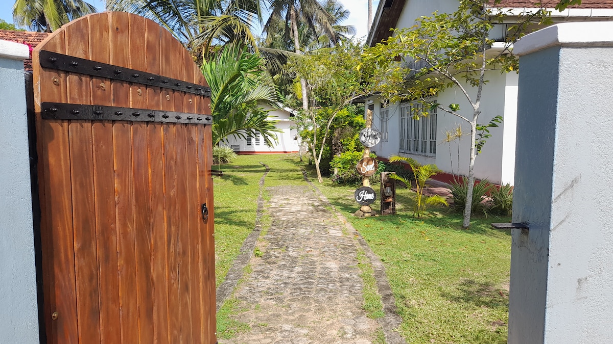 A wooden gate opens to a charming pathway lined with lush greenery. The cobblestone path leads through a well-kept garden, flanked by palm trees and foliage, towards a welcoming entrance of the property.