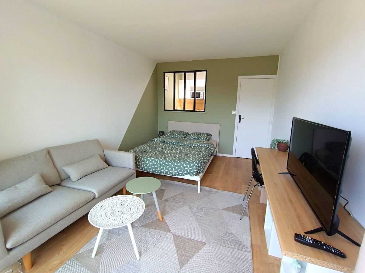 A modern living space features a two-seat sofa and a spacious bed with patterned bedding. A flat-screen TV is mounted on a wooden media unit beside a light circular table. Natural light enters through a large window, enhancing the room's bright atmosphere.