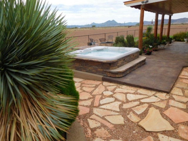 A spacious outdoor area is seen, featuring a hot tub surrounded by a stone barrier and mature landscaping. A paved walkway leads to the hot tub, complemented by a variety of plants. In the distance, a rural landscape with low mountains is visible.