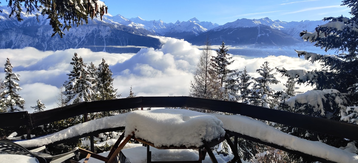 A balcony with a snowy railing overlooks a sea of clouds and majestic mountain peaks. Snow-covered trees frame the scene, enhancing the serene atmosphere of the Alps under a clear blue sky.
