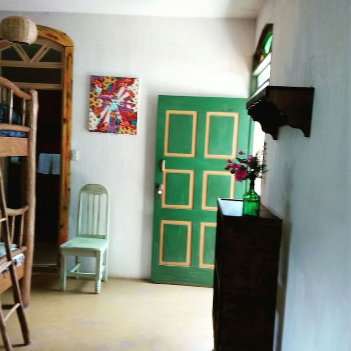 La litera, dos camas más. Una repisa con perchas y un mueble para acomodar ropa. Una silla que es siempre útil tener en una habitación. La entrada al baño, la puerta verde de entrada a la habitación.