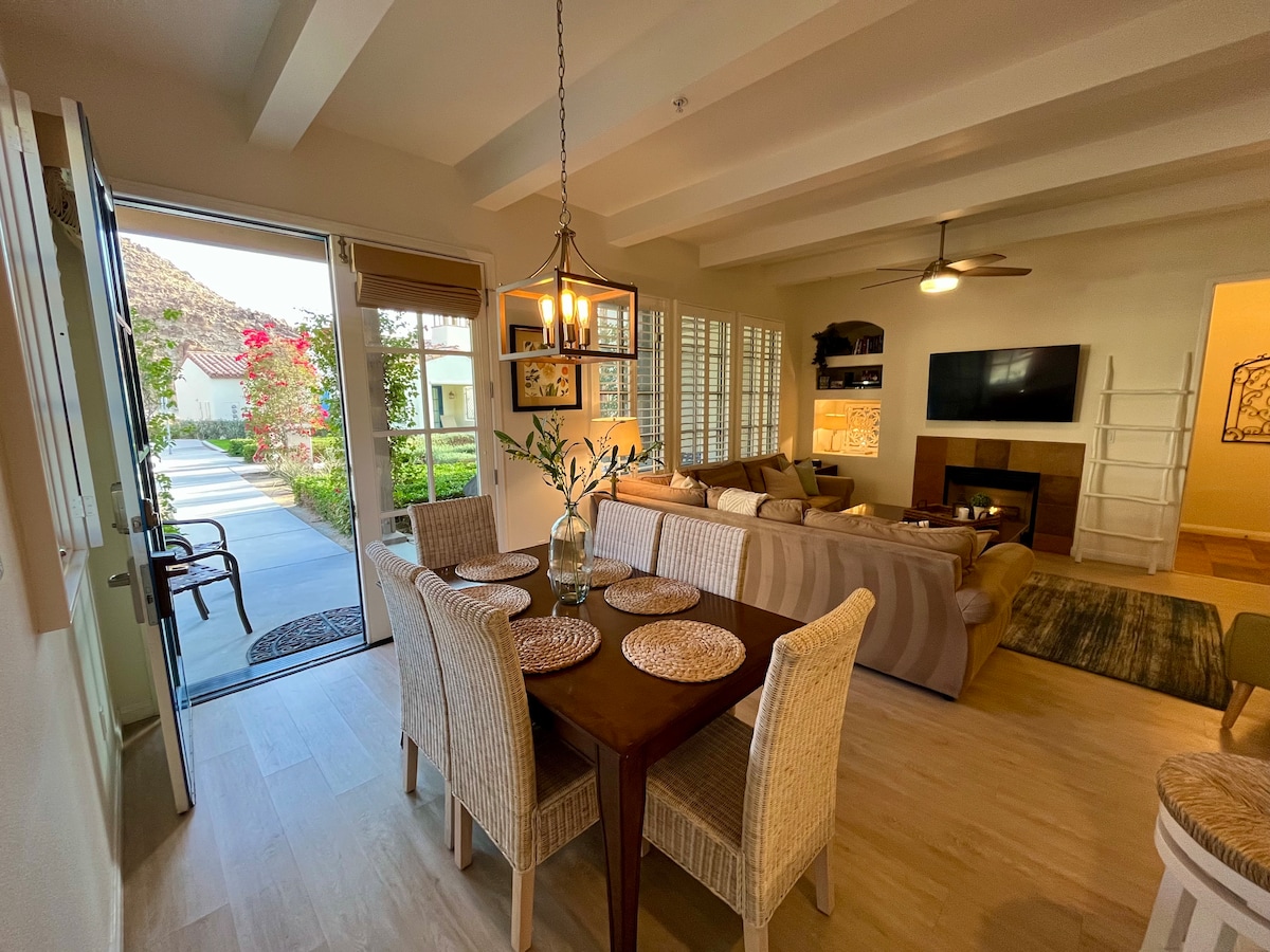 A spacious living area is visible, featuring a comfortable sofa arranged around a coffee table. A dining table with woven placemats sits beside a door leading to an outdoor area. Natural light illuminates the room, highlighting the elegant decor and inviting ambiance.