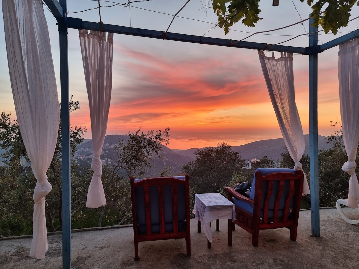 A serene outdoor seating area features two wooden chairs with comfortable cushions, positioned on a patio overlooking a vibrant sunset. Curtains sway gently in the breeze, framing the picturesque view of rolling hills and the sea in the distance.