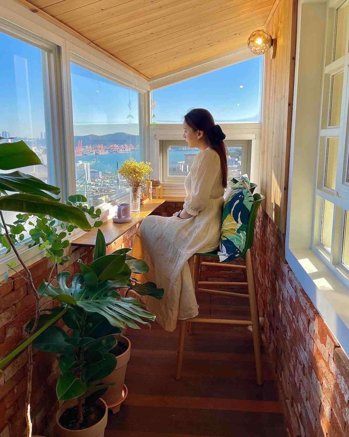 A serene corner of the room features large windows offering a view of the landscape. A wooden counter is accompanied by a tall stool, with potted plants adding a touch of greenery. Natural light enhances the warm tones of the wooden walls.