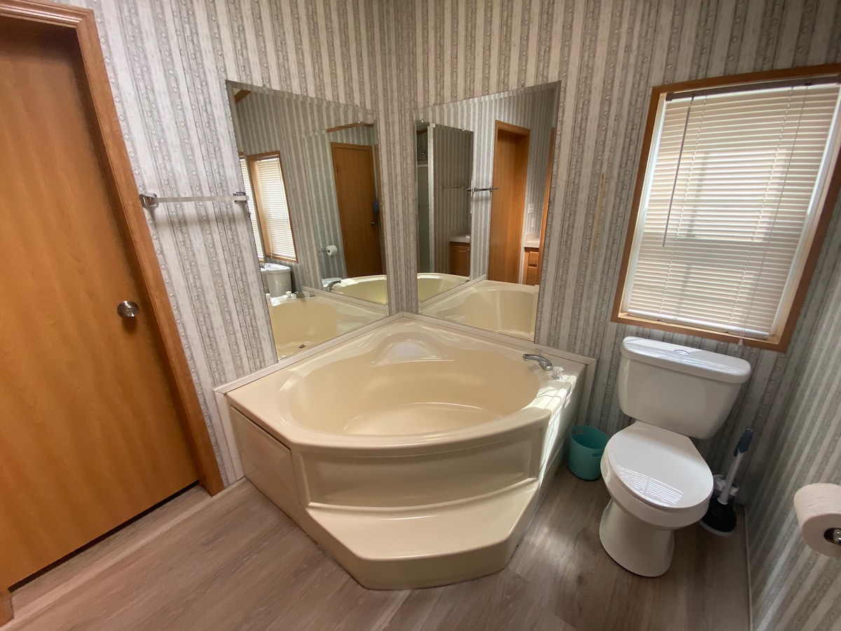 A well-appointed bathroom features a spacious corner bathtub, complemented by three mirrors that enhance the sense of space. A toilet is positioned nearby, with natural light filtering through a window adorned with slatted blinds.