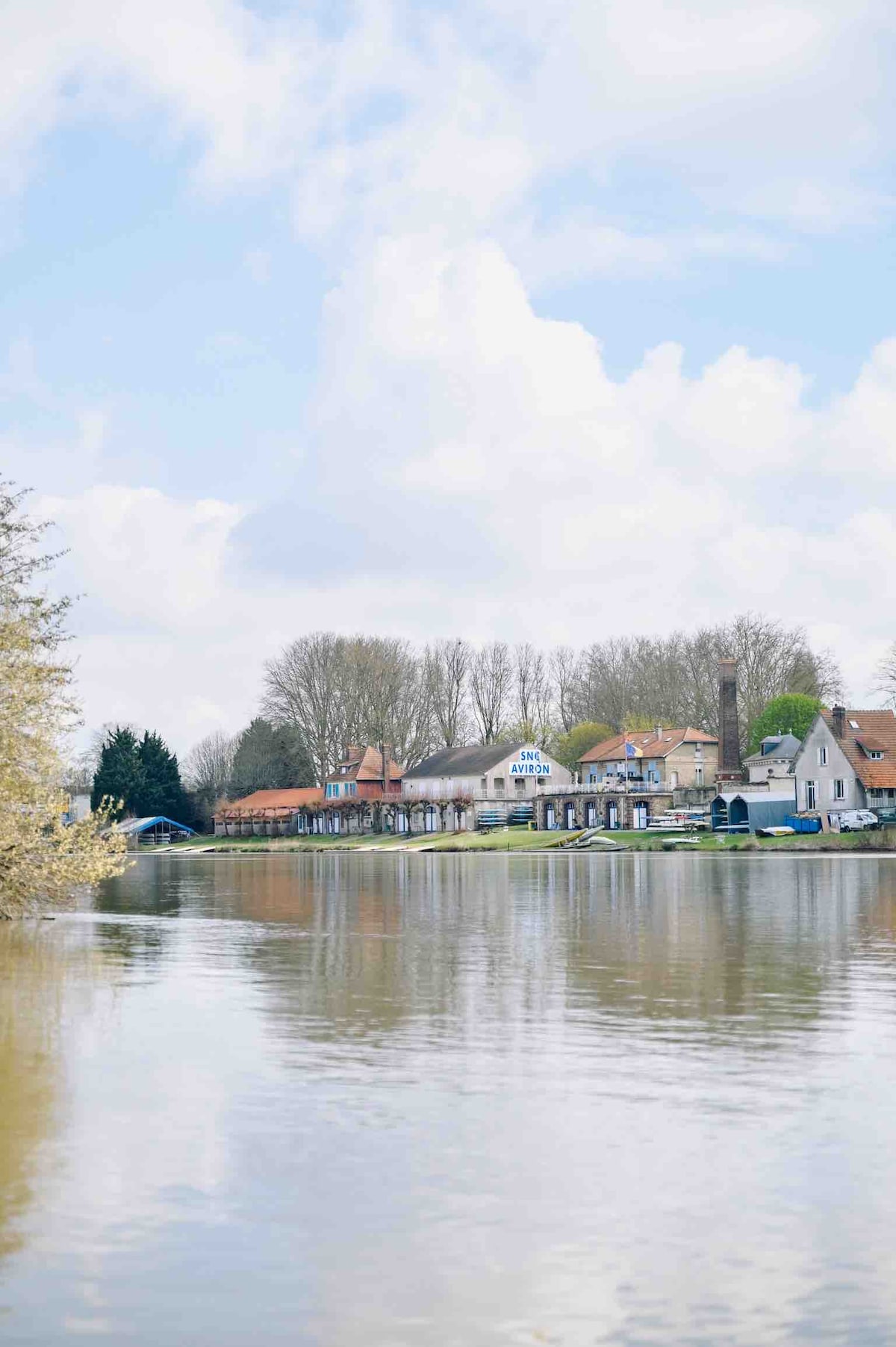 The image captures a serene waterfront view, showcasing a row of charming buildings along the riverbank. The calm water reflects the soft clouds overhead and the lush trees lining the shore, providing a tranquil atmosphere.