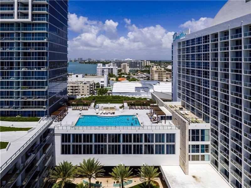 An aerial view of the property is presented, showcasing a large swimming pool surrounded by lounge chairs. The pool area is bordered by well-maintained landscaping, including palm trees. The backdrop features high-rise buildings and a glimpse of the Miami coastline under a partly cloudy sky.