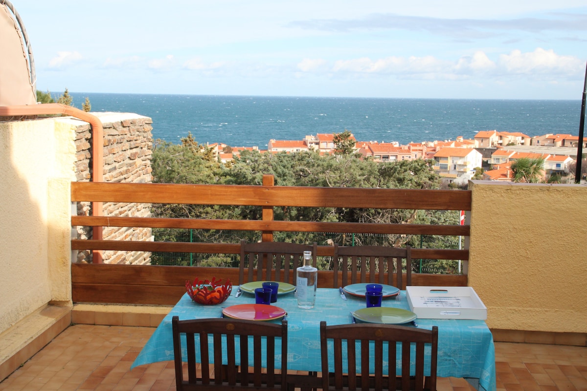 A dining table with colorful plates and glasses is set on a terrace overlooking the sea. The structure features wooden railings, and the coastline is visible in the background, showcasing a blend of greenery and rooftops. The sky is partly cloudy, adding to the scenery.