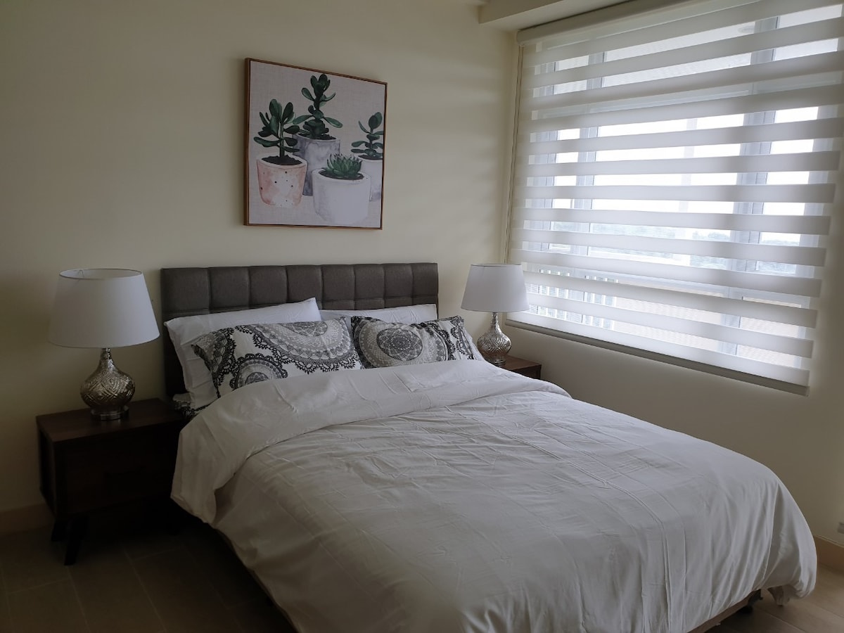 A contemporary bedroom features a spacious bed adorned with a light-colored duvet and patterned pillows. The room is complemented by two bedside tables with matching lamps. Large windows with sheer and solid blinds allow natural light to filter in, enhancing the serene atmosphere.