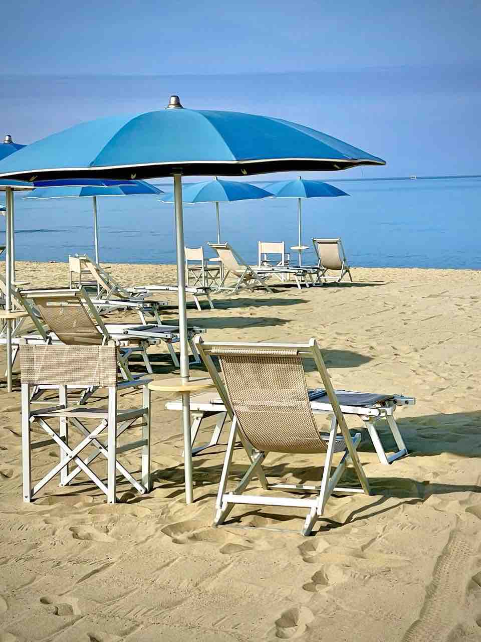 A sandy beach scene is displayed, featuring several sunbeds under blue umbrellas. The calm water glistens in the background, with a clear horizon meeting the sky. The arrangement of sunbeds provides ample space for relaxation, inviting visitors to enjoy a day by the sea.