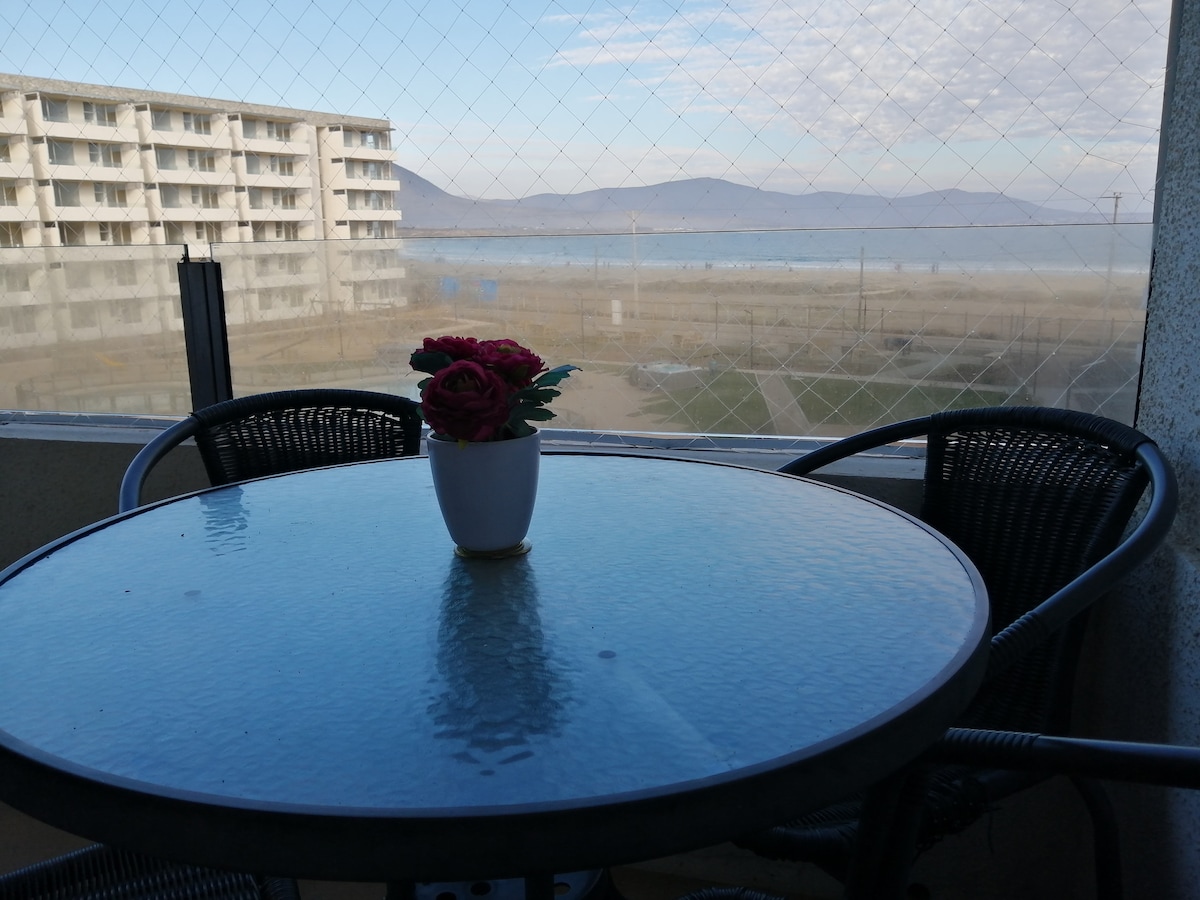 A round glass table is set with a small potted flower, providing a serene view of the beach and mountains in the background. The scene is framed by a mesh screen, indicating a spacious balcony overlooking the tranquil waters.
